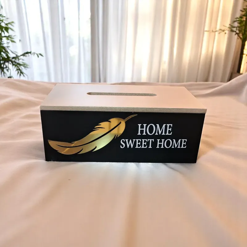 صندوق مناديل خشبي أنيق باللونين الأسود والعسلي، مزين برسم ذهبي على شكل ريشة وعبارة "HOME SWEET HOME". مثالي لتزيين المنزل وتنظيم المناديل.