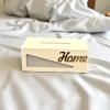علبة مناديل خشبية فاخرة بلون بيج مع كلمة "Home" ذهبية، موضوعة على غطاء سرير