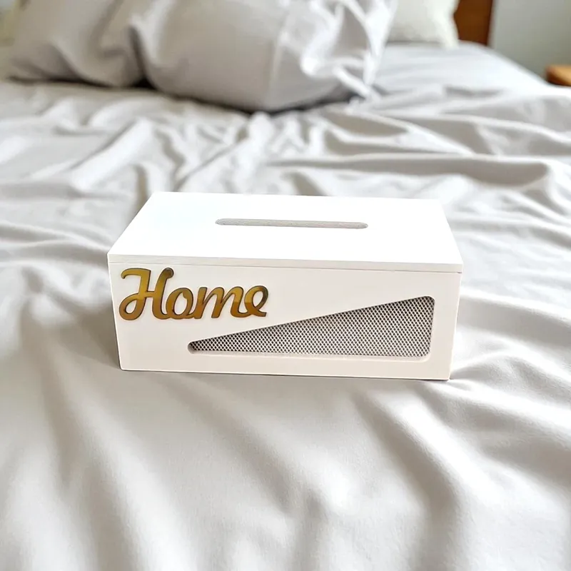 علبة مناديل خشبية باللون الأبيض مع كلمة "Home" ذهبية بارزة، موضوعة على سرير كقطعة ديكور منزلية