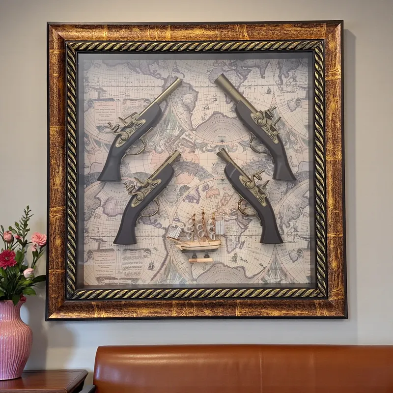 لوحة حائط ديكور مربع بإطار ذهبي وبني، تحتوي على 4 مسدسات عتيقة ومجسم سفينة شراعية على خلفية خريطة عالم قديمة.