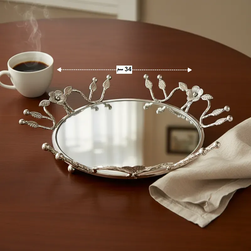 طقم صواني تقديم دائري فاخر بتصميم زهور وفروع، فضي (قطعتان)، موديل 030397 صينية فضية بيضاوية عاكسة صغيرة الحجم (34 سم) مزخرفة بالزهور، بجانب فنجان قهوة ساخن على طاولة خشبية.