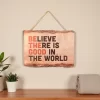 لوحة جدارية خشبية معلقة مكتوب عليها BELIEVE THERE IS GOOD IN THE WORLD، تصميم ريفي وألوان برتقالية وبنية، ديكور حائط إيجابي.