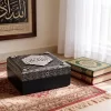 صندوق حفظ مصحف إسلامي فاخر بنقوش فضية كريستالية وغطاء أسود وجسم أزرق داكن مغلق وموضوع بجانب مصاحف.