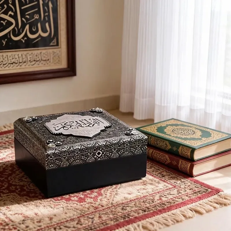 طقم إسلامي فاخر: صندوق حفظ المصحف الشريف مع سبحة، موديل 030582 صندوق حفظ مصحف إسلامي فاخر بنقوش فضية كريستالية وغطاء أسود وجسم أزرق داكن مغلق وموضوع بجانب مصاحف.
