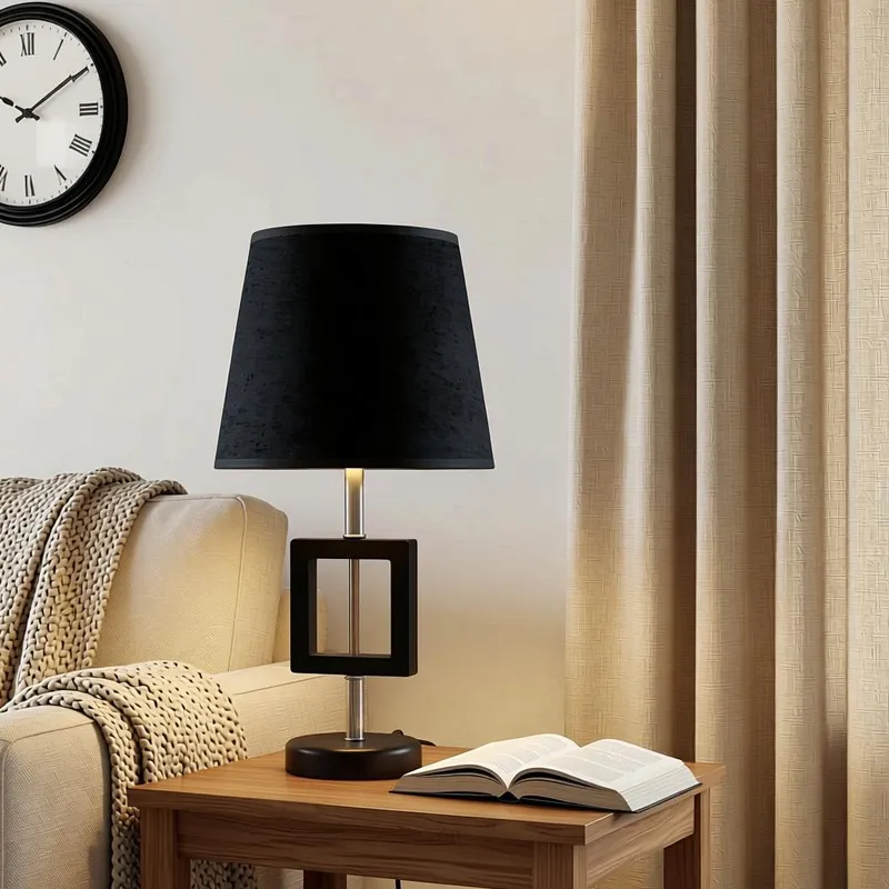 اباجورة مودرن سوداء بحامل معدني هندسي، موديل 028721، ارتفاع 46 سم اباجورة مودرن بجزء خشبي أسود مفرغ وشابوه قماش مكملة لديكور غرفة معيشة دافئة بجانب ستائر كريمي وكتاب مفتوح.