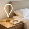 أباجورة ليد ذهبية تصميم دمعة مضاءة بضوء دافئ فوق كومودينو بجانب السرير مع كتاب ونظارة في غرفة نوم مودرن.