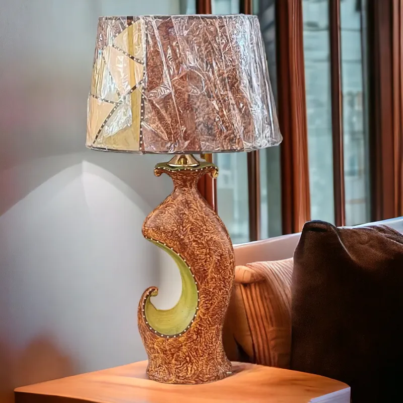 أباجورة خزفية بتصميم فني مبتكر يجمع بين الأشكال العضوية والألوان الدافئة. قاعدة الأباجورة على شكل إناء منحوت بتصميم فني مميز، مما يضفي عليها طابعًا فنيًا وجذابًا. وتتميز بمزيج من الألوان المتناسقة لكل من الغطاء والقاعدة.