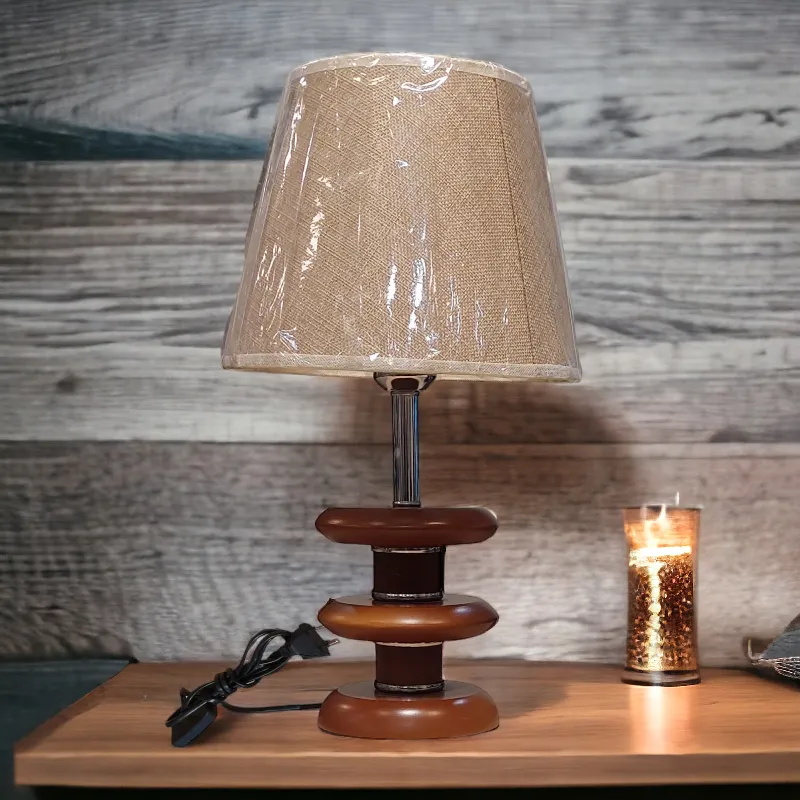 أباجورة نوم خشبية حديثة تتكون من قاعدة خشبية دائرية الشكل تتكون من ثلاث طبقات متداخلة، مما يعطي المصباح مظهرًا عصريًا. يرتكز على القاعدة ساق معدنية رفيعة تحمل الغطاء. الغطاء مصنوع من قماش ناعم بلون بيج فاتح، مما يوفر إضاءة دافئة ورقيقة.