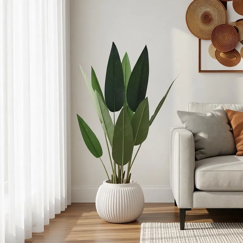 شجرة عصفور الجنة صناعية خضراء بأوراق جلدية في أصيص أبيض مضلع بجانب أريكة رمادية وديكور جداري خشبي.
